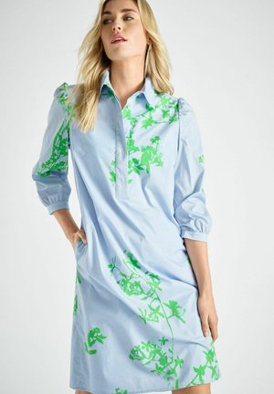 Vrouw in een lichtblauwe jurk met groene bloemenprint, staand met één hand in haar zak tegen een effen achtergrond.