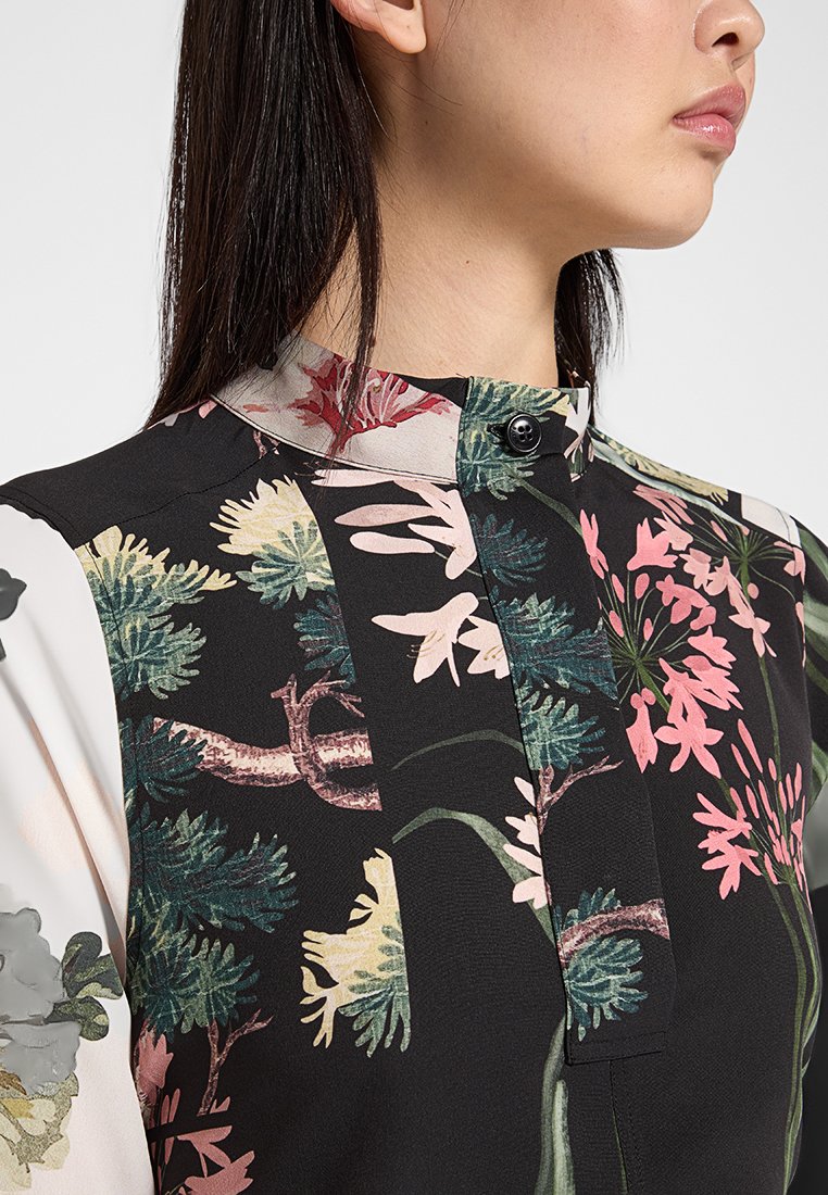 Zwarte blouse met bloemenpatronen in roze, groen en beige. Voorzien van een opstaande kraag en een knoopsluiting aan de voorkant. Gelaagde stof.