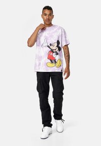 Fialové tričko s vířivým vzorem a grafikou Mickeyho Mouse v červených kraťasech. Nosič má na sobě černé cargo kalhoty a bílé tenisky.