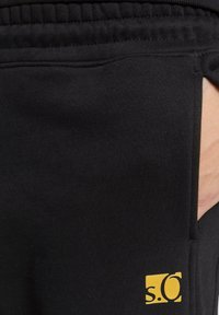 Schwarze Sweatpants mit elastischem Bund, ausgestattet mit einer seitlichen Tasche und einem kleinen gelben Logo-Akzent auf der unteren linken Seite. Weicher Stoff.