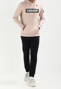 Felpa rosa chiaro con grande logo nero "CHASIN'", tasca a marsupio e pantaloni jogger neri. Indossata con sneakers grigie e bianche.