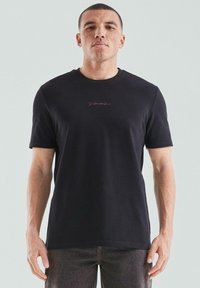 Schwarzes T-Shirt mit kurzen Ärmeln aus weichem Material, mit einem dezenten pinken Logo auf der Brust. Einfaches, klassisches Design mit rundem Ausschnitt.