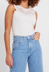 Kvinna klädd i en ärmlös topp med vit spetskant, instoppad i höga ljusblå jeans, med ena handen i fickan.