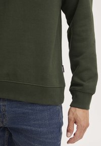 Olijfgroene sweatshirt met een geribde onderrand en manchetten. De stof ziet er zacht uit. Heeft een klein merkje aan de ondernaad. Draag het met een blauwe spijkerbroek.