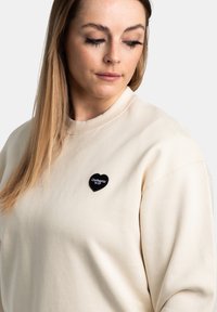 Beige sweatshirt met een ronde halslijn, voorzien van een zwart hartvormig geborduurd merkteken met de tekst "Carhartt WIP." Soepele stoftextuur.
