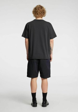 Black cotton t-shirt with short sleeves, back view, paired with navy blue shorts and black shoes, showing a relaxed fit and simple design.