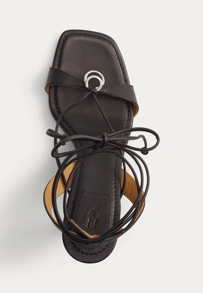 Μαύρο δερμάτινο σανδάλι με επίπεδη σόλα και τετράγωνη μύτη, ασημένια κυκλική αγκράφα και μακριές λουριά που τυλίγονται γύρω από τον αστράγαλο σε λευκό φόντο.
