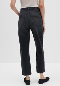 Persoon in hoge taille zwarte straight-leg jeans en zwarte ballerina's, staand tegen een effen lichtgrijze achtergrond.