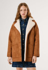 Cappotto in suede marrone con colletto in shearling bianco, vestibilità oversize, con tasche anteriori e un vestito in denim a bottoni sotto.