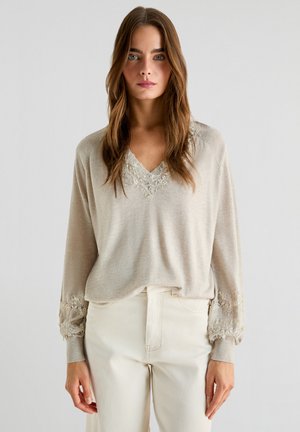 Beige dlouhý rukáv top s výstřihem do V a jemnou krajkovou výzdobou kolem výstřihu a manžet, spárovaný s světlými, fitted kalhotami.