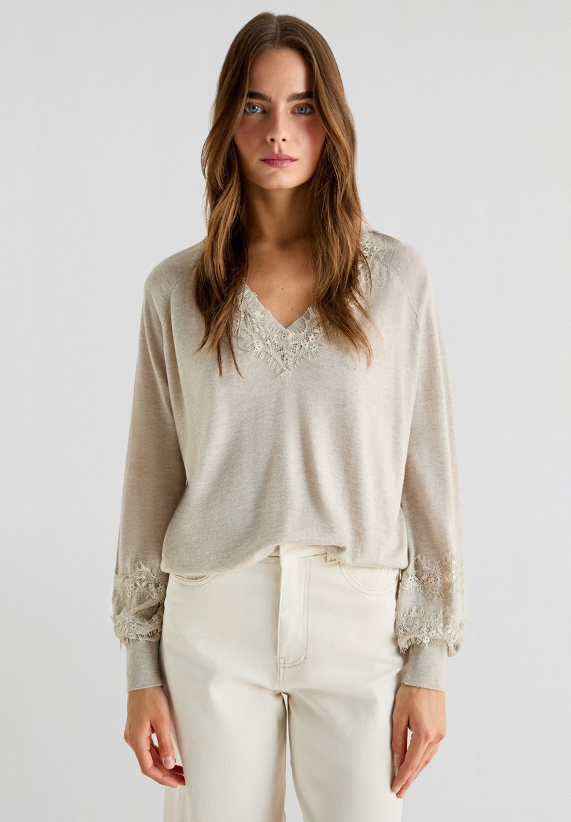 Maglietta beige a maniche lunghe con scollo a V e delicati inserti in pizzo attorno al collo e ai polsini, abbinata a pantaloni attillati di colore chiaro.