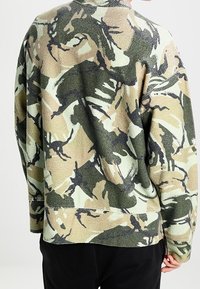 Persona vistiendo una chaqueta de forro polar de manga larga con un patrón de camuflaje abstracto en beige, verde y negro, vista desde atrás.
