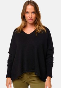 Schwarzer V-Ausschnitt-Pullover mit lockerer, oversized Passform, gerippter Textur und herunterhängenden Ärmeln. Kombiniert mit olivegrünen Hosen.