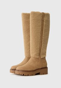 Botas hasta la rodilla con una sección inferior de ante color marrón claro y una parte superior de borrego esponjoso en tono marrón claro. Suela de goma con un patrón destacado para mayor tracción.