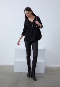 Black zippered top with three-quarter sleeves, paired with high-shine black leggings and ankle boots. Model poses against a white background.
