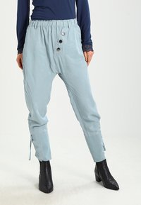 Světle modré kalhoty se širokým střihem, elastickým pasem, třemi ozdobnými knoflíky a šňůrkami na kotnících, doplněné černými lodičkami na podpatku.