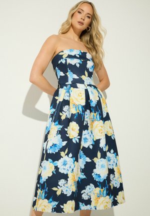 Femme portant une robe bustier bleu marine avec un grand motif floral jaune et bleu, debout devant un fond clair.
