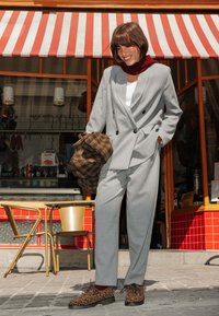 Costume gris à rayures avec blazer croisé et pantalon ajusté. Sac à carreaux marron et chaussures à imprimé léopard. Haut blanc et écharpe rouge.