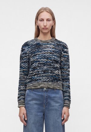 Femme aux cheveux blonds raides porte un pull en maille à motifs bleus, noirs et blancs et un jean bleu taille haute sur un fond uni.