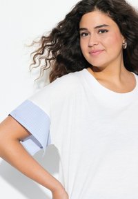 Valkoinen T-paita, jossa on lyhyet siniraidalliset hihat, pehmeä kangas, väljät mitat, pyöreä kaula-aukko ja hienovarainen tekstuuri vartalossa.