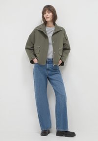 Femme portant une veste verte olive, un shirt gris, un jean bleu large et des bottes noires, se tenant contre un fond blanc avec les mains dans les poches.