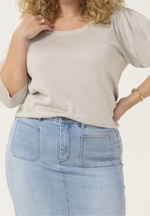 Kvinde iført en beige skinnende bluse med puffede ærmer, der er stoppet ned i et lyseblåt denimsnørre med forlommer.