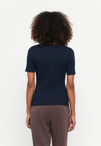 Άτομο με σγουρά μαλλιά που φοράει μπλε κοντομάνικο μπλουζάκι και καφέ παντελόνι, στέκεται με πλάτη σε λευκό φόντο.