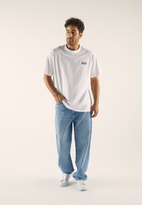 White cotton T-shirt with short sleeves and a small graphic on the left chest, paired with loose-fitting light blue jeans and white sneakers.