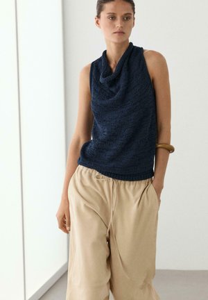 Vrouw draagt een mouwloze marineblauwe top met watervalhals en losse beige broek, handen in de zakken, staand tegen een effen lichte achtergrond.