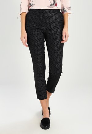 Femme portant un pantalon court texturé noir et des chaussures noires à enfiler avec des pompons, associées à un haut rose clair à motifs d’oiseaux.