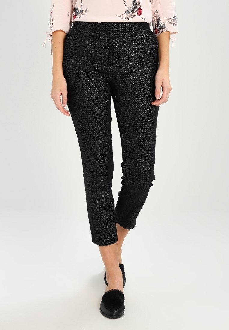 Femme portant un pantalon court texturé noir et des chaussures noires à enfiler avec des pompons, associées à un haut rose clair à motifs d’oiseaux.