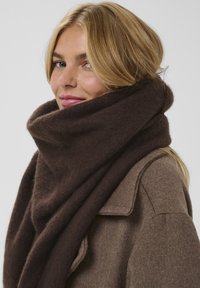 Brun uld tørklæde draperet omkring halsen, med en blød, tekstureret overflade. Det har et chunky, oversized design, der komplementerer en taupe frakke.