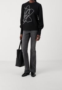 Schwarzer Strickpullover mit weißem Blumenranken-Design, kombiniert mit grauen ausgestellten Jeans und schwarzen Stiefeletten aus Lackleder, dazu eine schwarze Tote-Tasche.