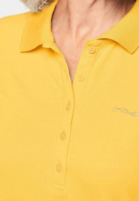 Geel polo shirt van katoen; heeft een gestructureerde afwerking, een drieknoopsluiting en een zilveren geborduurd design op de borst.