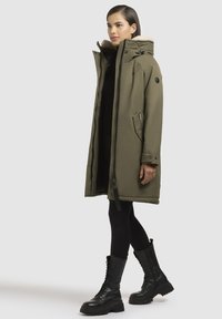 Olijfgroene lange parka met een zachte bontvoering in de capuchon, ritssluiting aan de voorkant, twee zijzakken en zwarte rubberen veterboots.