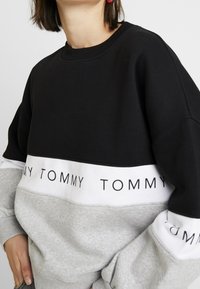 Sudadera negra y gris con un diseño voluminoso. Presenta una banda blanca con el texto "TOMMY" repetido. Tejido suave con cuello y puños acanalados.