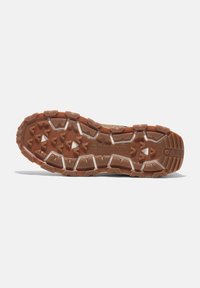 Suela de zapatilla de trail con un caucho marrón rugoso y texturizado, que presenta clavos triangulares pronunciados para una mejor tracción. Detalles en blanco que acentúan el diseño.