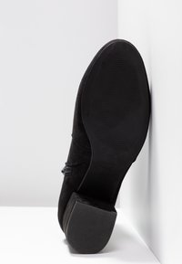 Botte en daim noir avec un petit talon carré vue du côté de la semelle, appuyée contre un angle blanc formé par un mur et le sol.