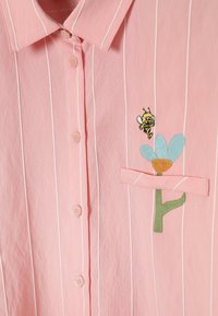 Rosa Hemd mit weißen vertikalen Streifen und einer kleinen bestickten Biene nahe einer hellblauen Blume auf der Brusttasche.