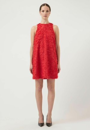 Kvinne stående, iført rød teksturert A-linje kjole uten ermer og svarte høyhælte sko, mot en ensfarget lys bakgrunn.