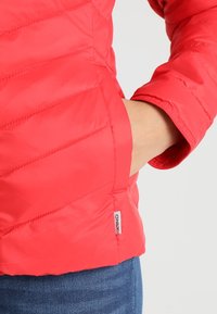 Rød pufjakke med diagonale syninger, der har en sidelomme og en logomærkat. Materialet er glat og skinnende med en tætsiddende pasform.