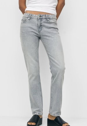 Personne portant un jean droit gris clair, un haut blanc et des chaussures noires à talons carrés ouverts à l'avant, debout devant un fond uni.