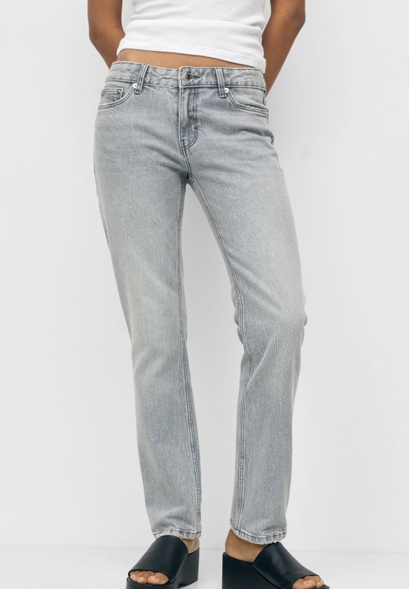 Personne portant un jean droit gris clair, un haut blanc et des chaussures noires à talons carrés ouverts à l'avant, debout devant un fond uni.