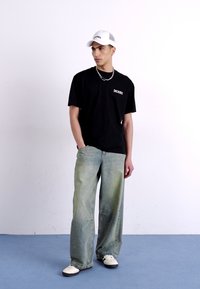 Camiseta negra de cuello redondo con logo blanco, jeans de pierna ancha azul claro, gorra blanca, collar de cadena plateado y zapatillas blancas.