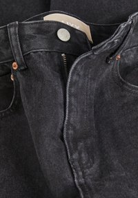 Svarta denimjeans med dragkedja, kopparnitar och en etikett inuti. Tyget har en något texturerad yta.