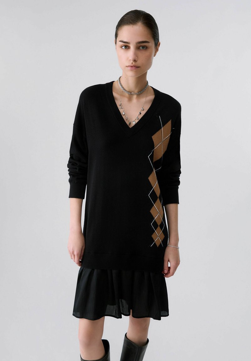 Νεαρή γυναίκα με μαύρο φόρεμα πουλόβερ V-neck με καφέ αργκίλ μοτίβο στη μία πλευρά, μαύρη φούστα και μαύρες μπότες, στέκεται μπροστά σε γκρι φόντο.