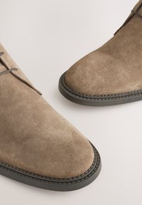 Hnedé semišové topánky s okrúhlou špičkou, sivými šnúrkami a kontrastnou čiernou gumovou podrážkou. Obsahujú šité detaily pozdĺž okraja podrážky.