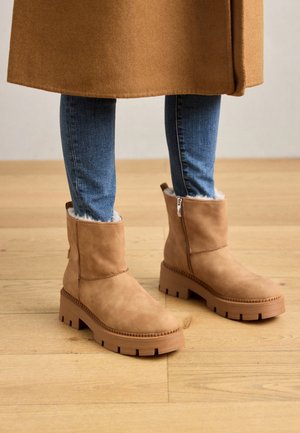 Kvinde iført blå skinny jeans og tanfarvede ruskindsstøvler med chunky såler og for i uld, stående på trægulv.