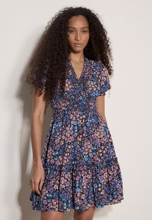 Kobieta o długich kręconych włosach, ubrana w sukienkę o krótkim rękawie, z dekoltem w serek, sięgającą do kolan, w kwiatowy wzór w odcieniach niebieskiego, fioletowego i różowego.