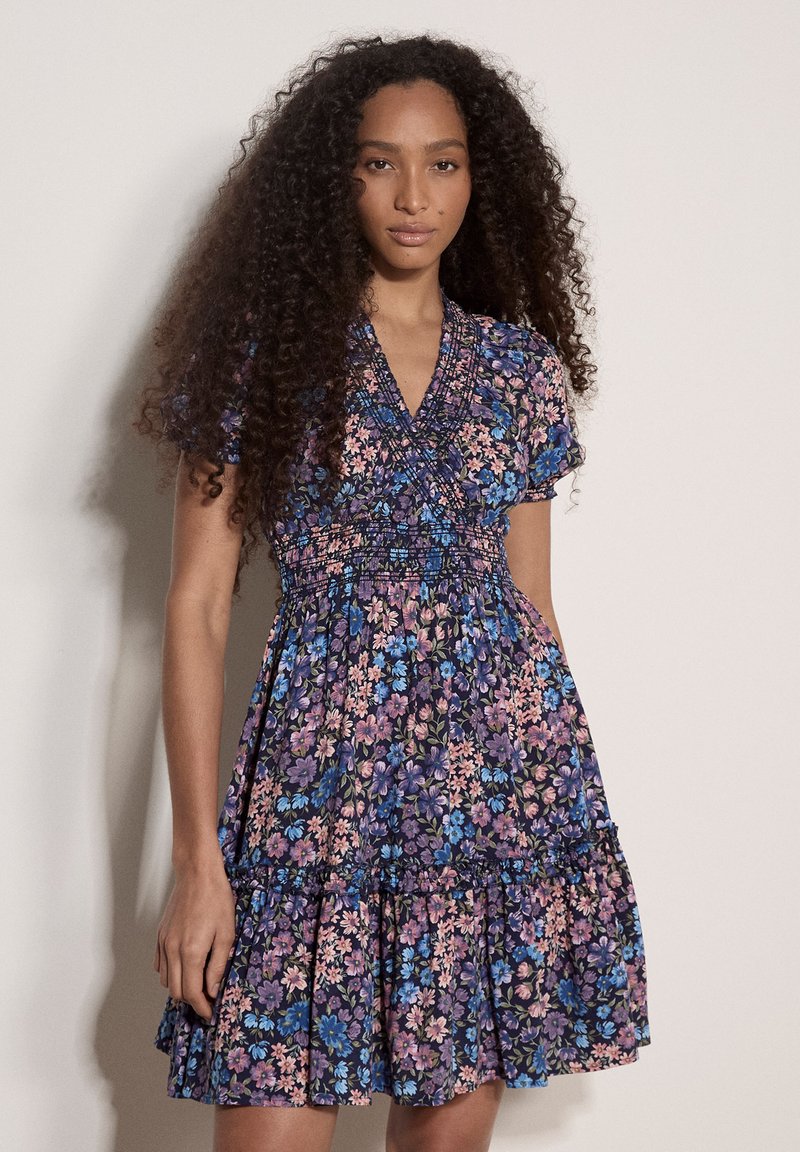 Femme aux longs cheveux bouclés portant une robe fleurie courte à manches courtes, col en V, longueur genou, dans des tons de bleu, violet et rose.